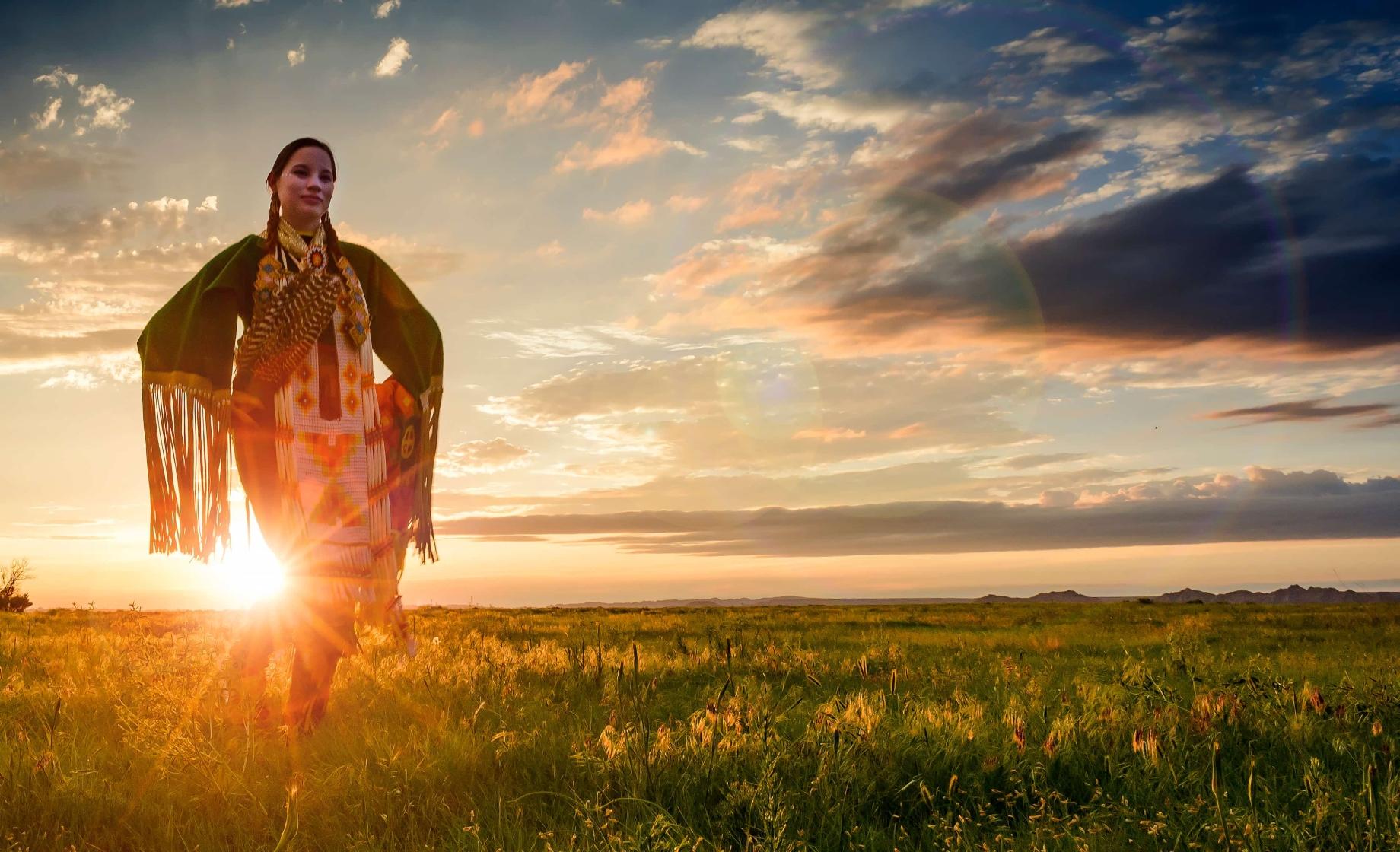 A Native American woman poses in regalia in front of a sunset on the Pine Ridge Reservation