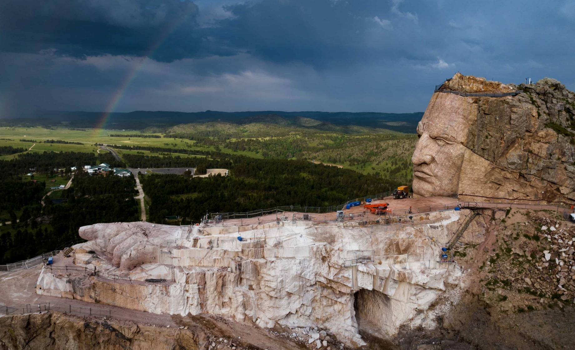 The face of the Crazy Horse sculpture looks out over an arm carving-in-progress with the green Black Hills forest and blue skies in the background